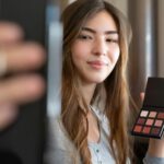 A young woman live streaming a makeup tutorial from her home office, using a smartphone.
